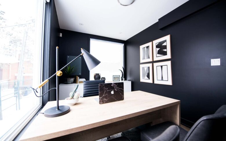 ufficio moderno con pareti nere, quadri bianchi e neri, scrivania in legno, lampada da tavolo in metallo, mac nero, pianta grassa e poltrona in pelle nera